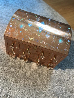 Charlotte Tilbury adventskalender - Säljer en exklusiv adventskalender från Charlotte Tilbury i skimrande kopparfärg med holografiska detaljer och små lådor med guldfärgade knoppar. Perfekt för att förvara skönhetsprodukter eller smycken. Lyxig och dekorativ design med stjärnor och magiska symboler.obs den är tom och inga produkter kommer med! 