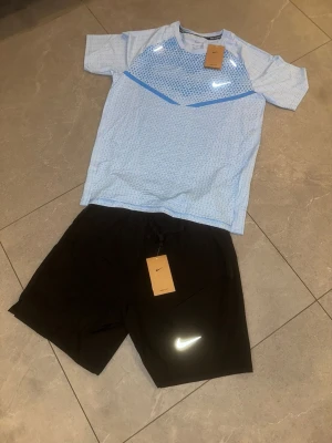 Nike träningsset ljusblå/svart - Snyggt träningsset från Nike med en ljusblå t-shirt med diskret mönster och reflekterande logga, samt svarta shorts med vit Nike-logga. Perfekt för löpning eller gym. Materialet är lätt och andas, vilket gör det bekvämt under intensiva pass.