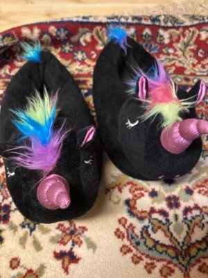Svarta enhörningstofflor med regnbågshår - Mysiga svarta tofflor formade som enhörningar med färgglatt regnbågshår och rosa horn i glansigt material. Tofflorna har broderade ögon och små öron, perfekta för dig som vill ha något lekfullt och unikt hemma. Materialet är mjukt plysch.