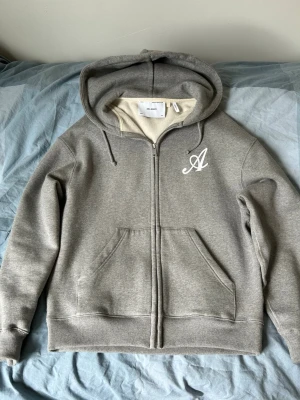 Axel arigato hoodie  - Snygg grå hoodie från Axel Arigato med hel dragkedja, huva med dragsko och stor ficka framtill. På bröstet sitter ett vitt broderat A som ger en clean look. Tillverkad i mjuk bomullsmix som känns skön mot huden. Perfekt för chill dagar eller när du vill vara casual.
