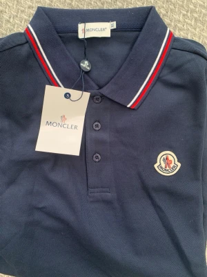 Marinblå pikétröja från Moncler - Snygg marinblå pikétröja från Moncler med klassisk krage och knappar framtill. Kragen har röda och vita ränder som ger en sportig touch. Tröjan är dekorerad med Monclers ikoniska logotyp på bröstet. Tillverkad i bomull för en bekväm känsla.
