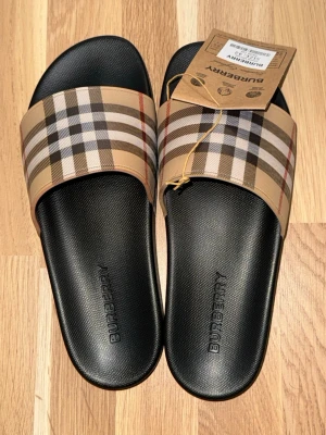 Burberry tofflor med klassiskt rutmönster - Säljer ett par snygga Burberry tofflor med det ikoniska beige, svarta och röda rutmönstret på remmen. Sulan är svart med präglad Burberry-logga. Perfekta för chill dagar hemma eller till stranden. Tillverkade i slitstark syntet.