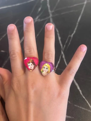 Två hjärtformade prinsessringar - Säljer två färgglada ringar med hjärtformade stenar, prydda med motiv av Disneyprinsessor. En ring är rosa med Belle från Skönheten och Odjuret, den andra är lila med Rapunzel. Båda ringarna har justerbar metallbas och är perfekta för dig som gillar Disney och vill ha något lekfullt.