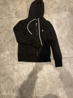 Svart hoodie från Polo Ralph Lauren - Snygg svart hoodie med dragkedja från Polo Ralph Lauren. Klassisk design med vit dragsko i huvan och den ikoniska loggan broderad på bröstet. Två fickor framtill och ribbade muddar. Perfekt för en avslappnad och stilren look.