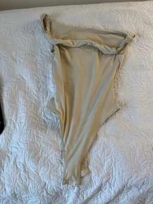 Beige off shoulder body - Trendig beige body med off shoulder-design och tryckknappar i grenen. Tillverkad i mjukt, stretchigt material som sitter snyggt mot kroppen. Perfekt att styla med jeans eller kjol för en clean look.