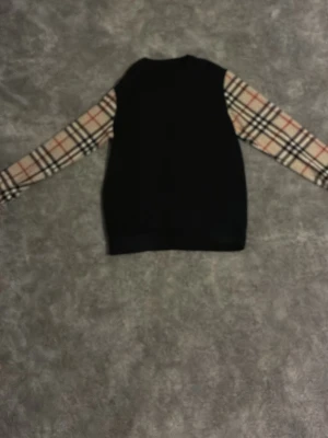 Svart skjorta med rutiga ärmar - Snygg svart skjorta med klassiska beige, röda och svarta rutiga ärmar. Skjortan har långa ärmar med knapp vid mudden och ger en stilren men ändå trendig look. Perfekt för dig som vill sticka ut lite extra.