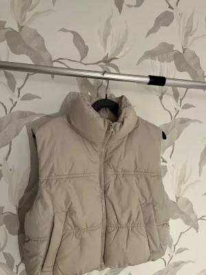 Beige puffig dunväst med hög krage - Snygg beige dunväst med puffig design och hög krage. Västen har dragkedja framtill och två fickor på sidorna. Perfekt för lager på lager och ger en trendig look. Materialet är quiltat och ger en mjuk känsla. Aldrig andvänt köpte och andvände den till slut inte har inte kvittot kvar för det var ett tag jag köpte den