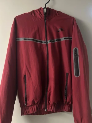 Vinröd Nike vindjacka med huva - Snygg vinröd vindjacka från Nike med svart dragkedja, reflexdetalj över bröstet och praktisk ficka på ärmen. Jackan har två fickor framtill, elastisk nederkant och huva. Perfekt för blåsiga dagar och sportiga outfits. Perfekt för sommaren