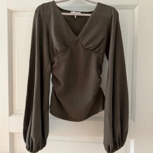 Olivgrön blus från NA-KD - Trendig olivgrön blus med djup v-ringning och långa ballongärmar. Tillverkad i ett lätt, strukturerat tyg som ger en snygg siluett. Perfekt att styla med både jeans och kjol för en cool look. Den är endast använd en gång och är därför i väldigt bra skick🤩