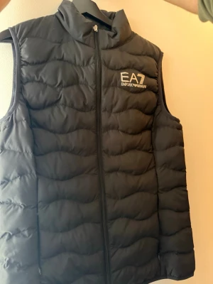 Svart dunväst från Armani - Snygg svart dunväst från Emporio Armani EA7 med hög krage och dragkedja framtill. Västen har ett stilrent, quiltat mönster och logga på bröstet. 