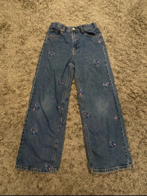 Blå jeans med Stitch-brodyr - Unika blå jeans med raka ben från Zara och har broderade Stitch-figurer samt små rosa blommor över hela byxorna. Jeansen har klassisk femficksmodell, knappgylf och resår baktill i midjan. Perfekta för dig som vill ha en lekfull och trendig stil.kostade över 600kr ny.