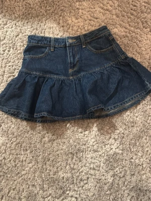 Mörkblå jeansskort med volang - Snygg mörkblå jeansskort med volangkant och klassiska fickor fram. Skorten har en feminin A-linjeform och är tillverkad i denim. Perfekt för dig som gillar en trendig och avslappnad stil.