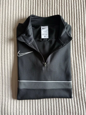 Svart Nike långärmad träningströja - Svart långärmad träningströja från Nike med halv dragkedja och vit logga på bröstet. Tröjan har en diskret vit linje och meshdetalj över bröstet. Perfekt för sport och träning, tillverkad i ett lätt och funktionellt material.