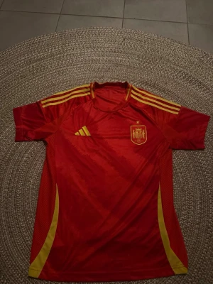 Spaniens matchtröja Adidas röd/gul - Säljer en röd matchtröja från Spaniens landslag med gula detaljer och Adidas-logga. Tröjan har korta ärmar, rund hals och tre gula ränder på axlarna. Tillverkad i lätt och ventilerande material, perfekt för fotboll eller träning.
