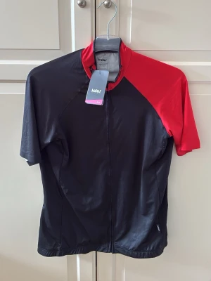 Svart och röd cykeltröja från Kalas - Snygg cykeltröja från Kalas med svart kropp och röd axel. Tröjan har korta ärmar, hel dragkedja framtill och är tillverkad i ett lätt och funktionellt material. Modellen är för kvinnor och har en sportig passform. Det står storlek 5, men passar som M/L. Helt ny!