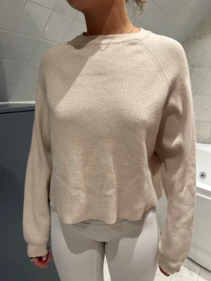 Beige stickad tröja från Pull & Bear - Säljer en beige stickad tröja med ribbad struktur och rund hals. Tröjan har lång ärm och en något croppad passform som ger en avslappnad vibe. Perfekt att matcha med jeans eller kjol för en enkel och snygg look.