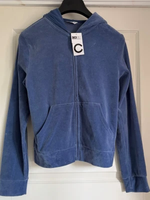 Blå velourhoodie från C - Mjuk och skön blå hoodie i velour från märket C. Hoodien har dragkedja framtill, två stora fickor och en klassisk huva. Perfekt för en chill och avslappnad stil. Tillverkad i bomullsmix med BCI-certifiering.
