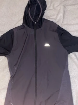 Svart vindjacka från Kalenji - Svart vindjacka från Kalenji med huva och hel dragkedja framtill. Tillverkad i lätt och funktionellt syntetmaterial som andas, perfekt för löpning eller annan träning. Reflekterande logga på bröstet och smidig passform.