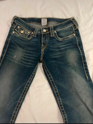 True Religion blå jeans med kontrastsömmar - Säljer ett par blå jeans från True Religion med tydliga kontrastsömmar och slitningar framtill. Klassisk femficksmodell med dekorativ bakficka och knappdetaljer. Jeansen har låg midja och är bootcut på mig. 