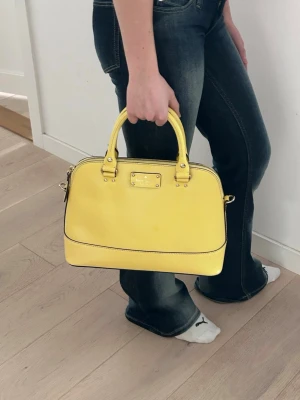 Gul handväska från Kate Spade - Snygg gul handväska från Kate Spade i slätt läder. Väskan har två handtag, guldfärgade detaljer och dragkedja upptill. Insidan är fodrad med mönstrat tyg och har innerfack för småsaker. Perfekt till våren och sommaren för dig som vill sticka ut med färg. Väskan är i nyskick.