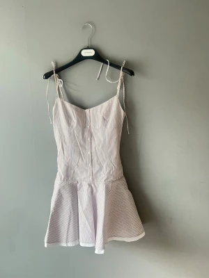 Randig kort klänning med knytband - Söt populär kort klänning med tunna axelband som knyts på axlarna. Klänningen är vit med tunna vertikala ränder i blått och har knappar framtill. Utsvängd kjol 