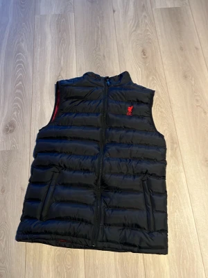 Svart dunväst från Liverpool FC - Ny pris 1099kr. Storlek S/M, jag är ungefär 172 och den passar mig. Jag köpte västen i Liverpool på deras butik precis bredvid arenan. 100% äkta alltså. Kontakta vid frågor eller fler bilder. Pris kan diskuteras.