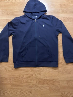 Mörkblå hoodie från Polo Ralph Lauren - Snygg mörkblå hoodie från Polo Ralph Lauren med dragkedja framtill och klassisk huva med dragsko. Tröjan har två fickor fram och den ikoniska vita Polo-loggan broderad på bröstet. Tillverkad i mjuk bomullsblandning för maximal komfort. Storlek XL men sitter mer som en L👍🏻skicka ett pris förslag 
