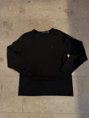 Svart sweatshirt Polo Ralph Lauren - Klassisk svart sweatshirt från Polo Ralph Lauren med rund halsringning och den ikoniska blå broderade loggan på bröstet. Tröjan har långa ärmar och ribbade muddar. Perfekt för en stilren och avslappnad look.