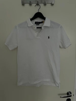 Vit pikétröja från Polo Ralph Lauren - Klassisk vit pikétröja från Polo Ralph Lauren med korta ärmar och den ikoniska broderade loggan på bröstet. Tröjan har krage och knappar vid halsen. Perfekt till sommaren och enkel att matcha med allt.