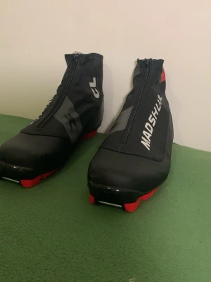 Madshus svarta kängor med dragkedja - Säljer ett par svarta kängor från Madshus med dragkedja framtill och röda detaljer på sulan. Skorna har sportig design, logga på sidan och är tillverkade i syntetmaterial som tål väta. Perfekta för dig som gillar en aktiv stil.