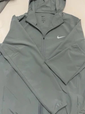 Grå vindjacka med huva från Nike - Snygg grå vindjacka från Nike med dragkedja framtill och huva. Jackan har en minimalistisk design med Nike-logga på bröstet och elastiska muddar vid ärmsluten. Perfekt för blåsiga dagar och enkel att matcha med olika outfits.