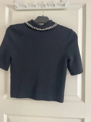 Svart topp med pärlkrage från H&M - Stilren svart topp från H&M med korta ärmar och en klassisk krage dekorerad med vita pärlor. Toppen har rak passform och är gjord i ett mjukt, stretchigt material som känns bekvämt mot huden. Perfekt för dig som vill ha en chic och trendig look.