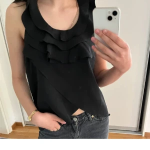 Svart ärmlös linnetopp med volanger - Snygg svart ärmlös topp med flera volanger framtill. Perfekt för både vardag och speciellare tillfällen. Den har en snygg och stilren design som passar till jeans eller kjol. Toppen är i ett mjukt och bekvämt material.