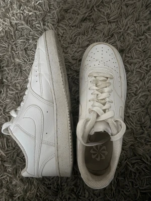 Nike Air Force 1 vita sneakers - Klassiska vita Nike Air Force 1 sneakers i lågt utförande. Tillverkade i läder med perforerad tåbox för ventilation och ikonisk swoosh på sidan. Tjock sula och vita skosnören ger en clean och tidlös look.