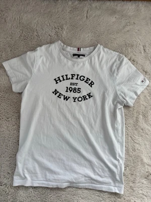 Vit Hilfiger t-shirt med tryck - Vit t-shirt från Tommy Hilfiger med svart texttryck 'Hilfiger Est. 1985 New York' på bröstet. Klassisk rund hals och korta ärmar. Liten Hilfiger-logga på ärmen. Tillverkad i mjuk bomull för en skön känsla.