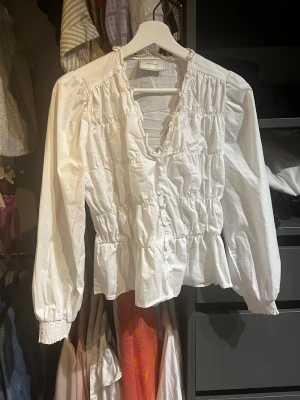 Vit blus med volanger från Neo Noir - Söt vit blus från Neo Noir med volangdetaljer och smock i midjan. Blusen har en djup v-ringning och långa puffärmar med resår vid ärmsluten. Tillverkad i ett lätt och luftigt tyg som ger en romantisk känsla.