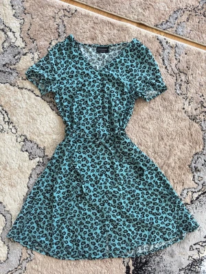 Turkos leopardmönstrad kortklänning - Kortklänning i turkos med svart leopardmönster. Klänningen har omlott framtill, korta ärmar och markerad midja för en smickrande siluett. Tillverkad i ett mjukt och lätt material som passar perfekt för varmare dagar. Anledning till att jag säljer är på grund av den är inte min stil längre. 