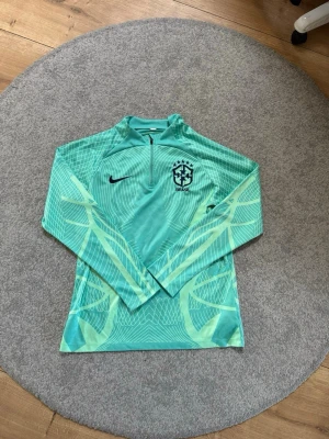 Nike Brasil långärmad träningströja - Snygg långärmad träningströja från Nike med Brasil-logga på bröstet. Tröjan är turkos med ljusgröna grafiska mönster, har halv dragkedja och hög krage. Tillverkad i ett lätt och funktionellt material som passar perfekt till fotbollsträning.