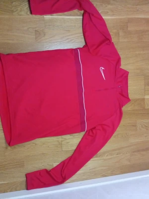 Röd långärmad träningströja Nike - Snygg röd långärmad träningströja från Nike med vit logga på bröstet och en tunn vit linje under bröstet. Tröjan har halv dragkedja framtill och är tillverkad i ett mjukt, stretchigt material som passar perfekt till träning eller uppvärmning.