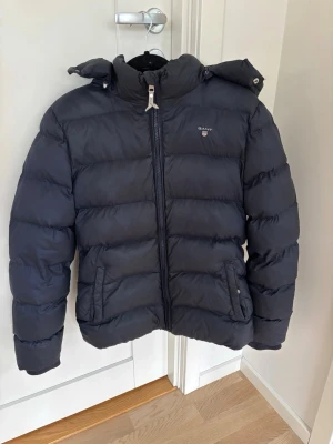 Mörkblå dunjacka från GANT - Snygg mörkblå (vinter) dunjacka från GANT med luva och dragkedja framtill. Jackan har en liten defekt på vänster arm, men den är lagad. Men det är absolut inget som syns över huvudtaget eller som man tänker på! Storlek 158/164 (13-14år) men skulle säga att den mer är storlek Xs/S. Den passar mig som är storlek S