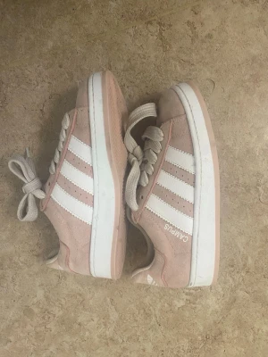 Adidas Campus rosa sneakers - Snygga Adidas Campus sneakers i ljusrosa mocka med vita klassiska ränder på sidorna och vit sula. Skorna har låga skaft, vita skosnören och en retrovibe som passar perfekt till streetwear. Adidas-logga och Campus-tryck på sidan.
