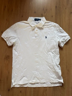 Vit pikétröja från Polo Ralph Lauren - Klassisk vit pikétröja från Polo Ralph Lauren med korta ärmar och krage i storlek M. Tröjan har två knappar framtill och loggan broderad i marinblått på bröstet. Pris kan diskuteras vid snabb affär.