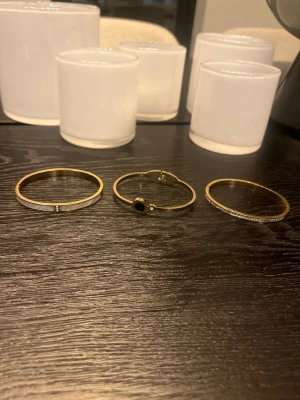 3X Guldiga armband med svart detalj - Säljer tre stilrena armband i guldfärgad metall. Ett av armbanden har en rund svart detalj och en liten gnistrande sten, medan de andra två är enkla och minimalistiska. Perfekta för att lyfta din outfit med en elegant touch. ALLA 3 för 279