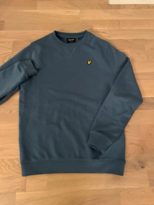 Blå sweatshirt från Lyle & Scott,helt ny utan prislapp har använts 1-2 gånger Max - Säljer en stilren blå sweatshirt från Lyle & Scott med rund halsringning och det klassiska gula logomärket broderat på bröstet. Tröjan har långa ärmar och ribbade muddar vid ärmslut och nederkant. Perfekt för en avslappnad look.