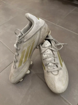 Vita Adidas fotbollsskor med guld - Säljer ett par vita fotbollsskor från Adidas med guldiga detaljer och klassiska tre ränder på sidan. Skorna har snörning, låg profil och dobbar för spel på gräs. Ovandelen är i syntetmaterial och designen är sportig och modern.