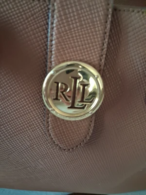 Brun handväska från Ralph Lauren deffekr - Snygg brun handväska från Ralph Lauren med guldfärgad RL-logga framtill. Väskan har två handtag och är gjord i ett strukturerat konstläder som ger en exklusiv känsla. Perfekt accessoar för att lyfta din outfit.