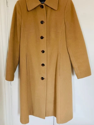 Kamelbrun ullkappa från Mexx - Stilren kamelbrun ullkappa från Mexx med klassisk krage och stora bruna knappar framtill. Kappan har rak passform, diskreta fickor i sidorna och är helfodrad. Perfekt för kyliga dagar när du vill ha en tidlös look.