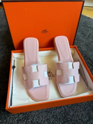 Hm oran sandeler i rosa  - Lyxiga Oran sandaler i ljusrosa läder med den ikoniska H-detaljen framtill. Sandalerna har öppen tå, platt sula och vita inslag på ovandelen. Perfekta för en trendig och avslappnad look. Levereras i originalförpackning.