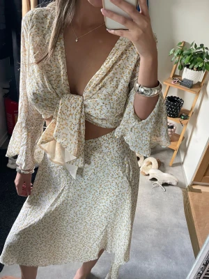 Blommig omlottklänning med knyt från Adoore  - Supersöt blommig omlottklänning i vitt och gult med stora knytband framtill och v-ringad urringning. 