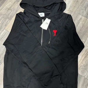 Svart hoodie från Ami med röd hjärtlogga - Svart hoodie från Ami med huva och half zip-dragkedja framtill. På bröstet sitter Ami:s ikoniska röda hjärta med bokstaven A broderat. Tröjan är i mjuk bomull och har ribbade muddar. Perfekt för dig som gillar stilrena detaljer och streetwear.